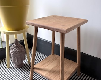 May include: A light brown wooden side table with a square top and a lower square shelf, supported by four straight legs. The table features visible wood grain and joinery details on the top surface, showcasing a natural finish.