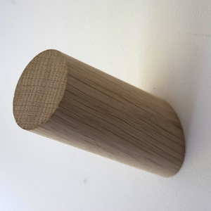 May include: A cylindrical wall hook crafted from light brown wood with a visible natural grain pattern. The hook features a flat circular front face and a smooth, elongated body that extends outward, providing a simple and functional design.