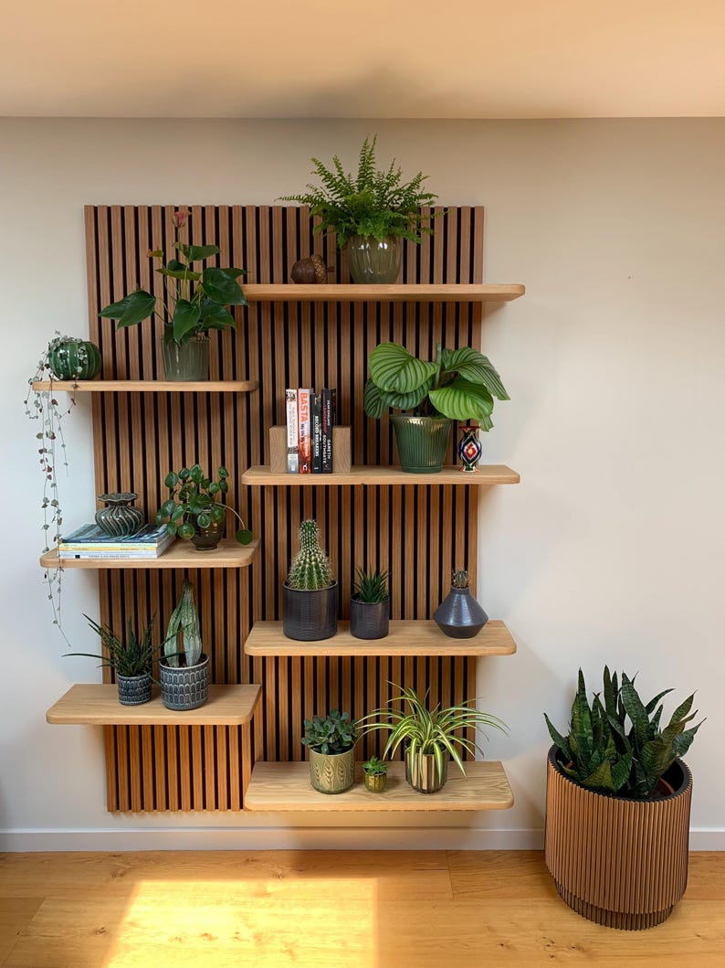 May include: A wall-mounted wooden slat panel featuring multiple light wood shelves holding various potted green plants, books, and decorative items. A large, cylindrical, slatted wooden planter with a snake plant sits on the floor.