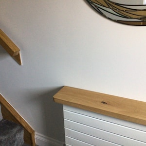 May include: A light oak wood radiator cover featuring a horizontal slatted white base and a natural wood grain top shelf, positioned next to a staircase with a matching oak handrail and a partial view of a decorative wall mirror.