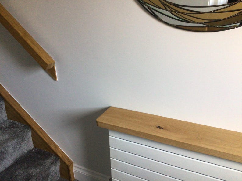 May include: A light oak wood radiator cover featuring a horizontal slatted white base and a natural wood grain top shelf, positioned next to a staircase with a matching oak handrail and a partial view of a decorative wall mirror.