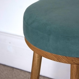 May include: A round stool featuring a dark teal velvet upholstered seat cushion. The base is constructed from light brown wood with a visible grain pattern, consisting of a circular frame and three sturdy, cylindrical wooden legs.