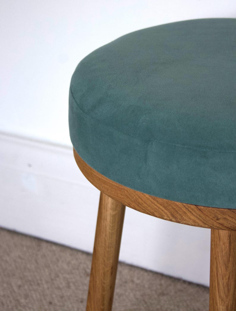 May include: A round stool featuring a dark teal velvet upholstered seat cushion. The base is constructed from light brown wood with a visible grain pattern, consisting of a circular frame and three sturdy, cylindrical wooden legs.