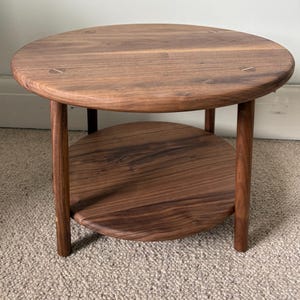 May include: A round, two-tiered side table made of dark brown wood. The top and lower shelf are circular, connected by three cylindrical legs. The table has a natural wood grain finish and measures approximately 20 inches in diameter.