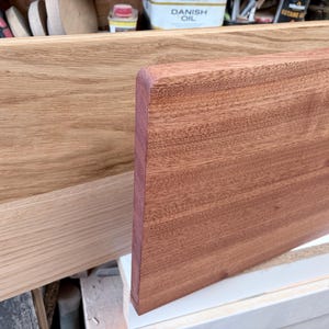 May include: Two rectangular wooden boards are displayed side by side. The board in the back is a light tan colour with a visible grain pattern. The board in the front is a darker reddish-brown colour with a smooth, rounded top edge.
