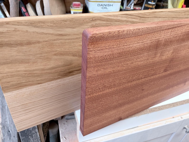 May include: Two rectangular wooden boards are displayed side by side. The board in the back is a light tan colour with a visible grain pattern. The board in the front is a darker reddish-brown colour with a smooth, rounded top edge.