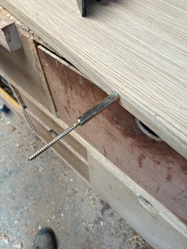 May include: A metal floating shelf support bracket with a threaded screw end, partially inserted into the edge of a light-coloured wooden board. The hardware features a cylindrical sleeve and a long, pointed, spiraled metal screw shaft.