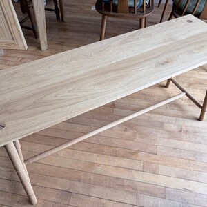 May include: A light-colored wooden bench featuring a rectangular top with visible grain patterns and four cylindrical legs. The top is secured with two circular wooden fasteners at each end, and a horizontal support bar connects the legs.