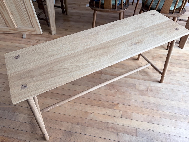 May include: A light-colored wooden bench featuring a rectangular top with visible grain patterns and four cylindrical legs. The top is secured with two circular wooden fasteners at each end, and a horizontal support bar connects the legs.