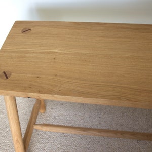 May include: A rectangular light brown wooden bench featuring a smooth grain surface. The top panel displays two circular joinery details with dark brown linear accents. The bench is supported by four cylindrical legs connected by a horizontal stretcher.