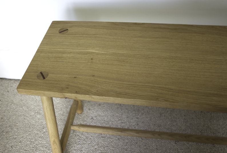 May include: A rectangular light brown wooden bench featuring a smooth grain surface. The top panel displays two circular joinery details with dark brown linear accents. The bench is supported by four cylindrical legs connected by a horizontal stretcher.