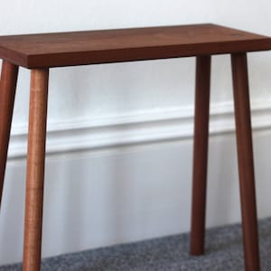 Könnte beinhalten: Ein kleiner Holzhocker mit einer rechteckigen Oberseite und vier Beinen. Der Hocker ist aus dunkelbraunem Holz gefertigt und hat ein schlichtes, minimalistisches Design.