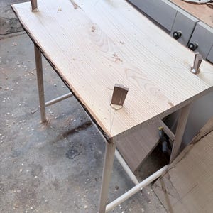 May include: A light-colored wooden table with a live edge and four legs. The tabletop is rectangular, with a natural, unfinished edge. The legs are thin and angled, with cross supports. Small wooden pegs are attached to the tabletop.
