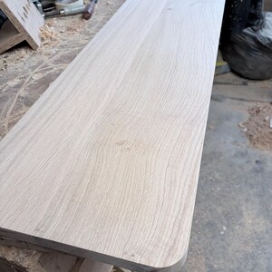 May include: A rectangular wooden board with a light tan colour and visible natural grain patterns. The board features a smooth, flat surface and one rounded corner, showcasing the raw texture of the wood material in a workshop setting.