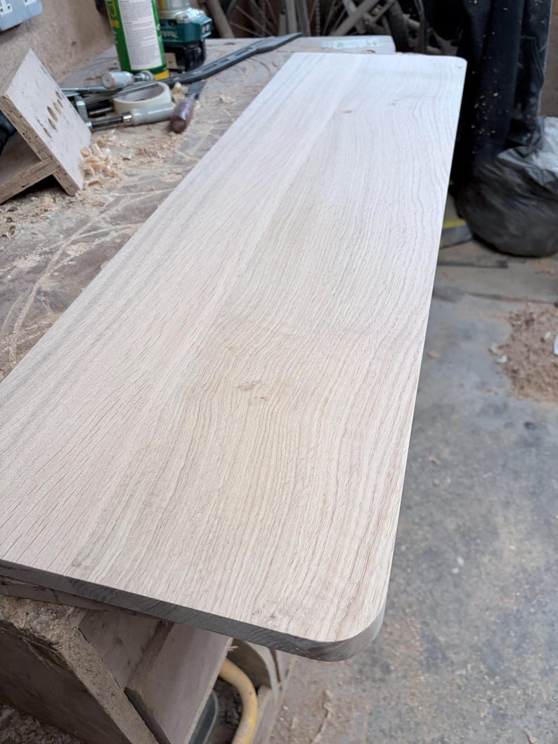 May include: A rectangular wooden board with a light tan colour and visible natural grain patterns. The board features a smooth, flat surface and one rounded corner, showcasing the raw texture of the wood material in a workshop setting.