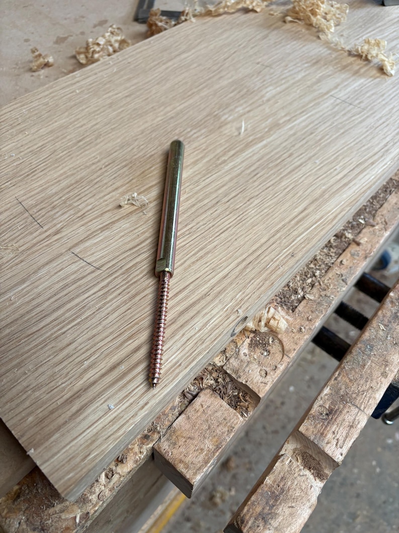 May include: A metal floating shelf bracket featuring a long, smooth cylindrical rod attached to a threaded screw base, resting on a light-coloured wooden board with visible wood grain and scattered wood shavings nearby.