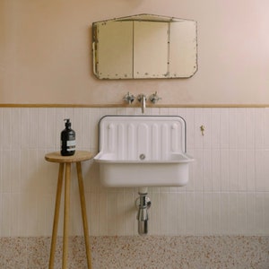 May include: A white porcelain sink with a black rim sits on a white tiled wall. A small wooden stool with a bottle of soap sits next to the sink. The sink is in a bathroom with a light pink wall and a small mirror above the sink.