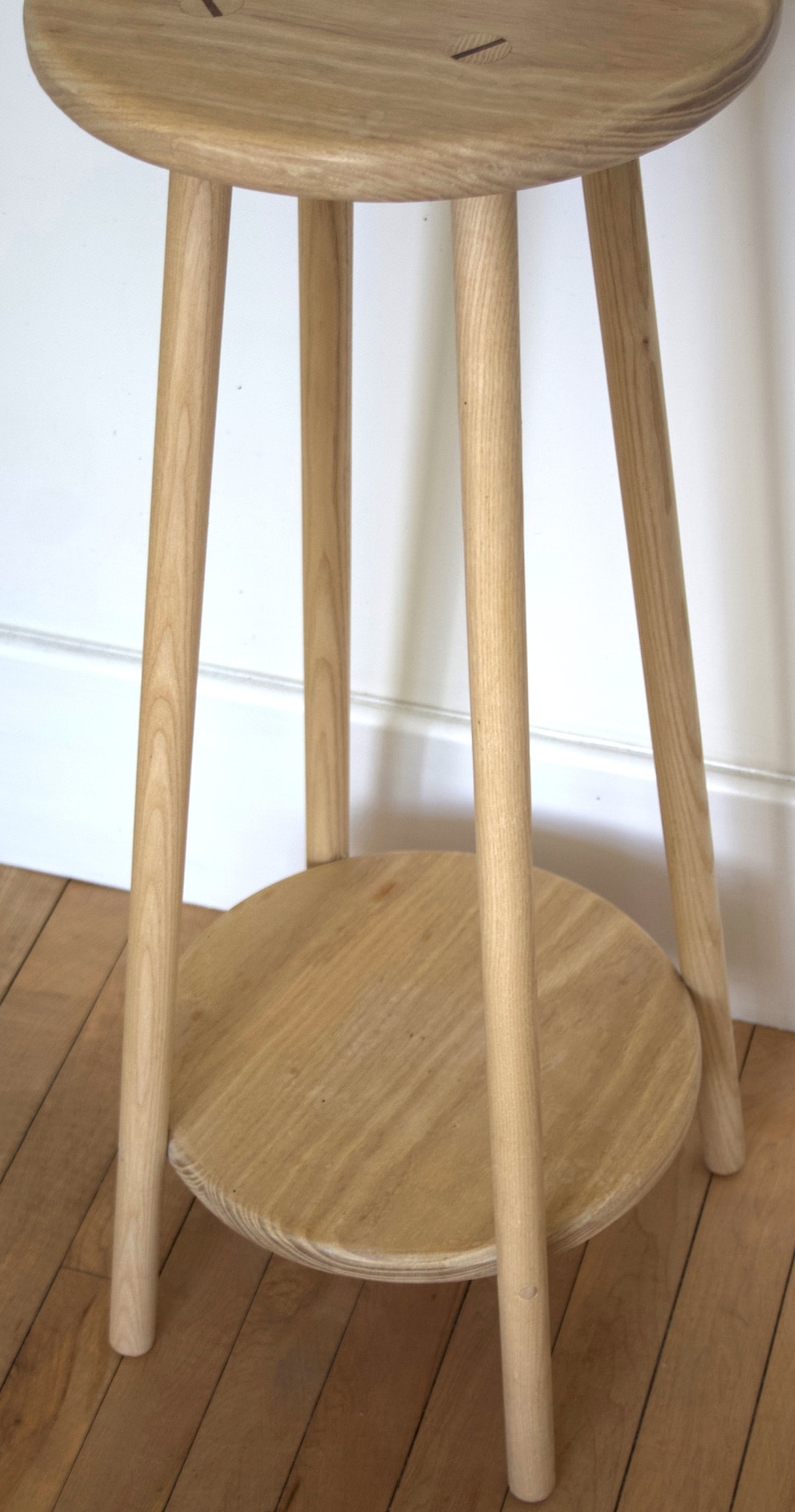 May include: A light wood side table with a round top and a lower shelf. The table has three legs and is made of natural wood.