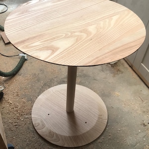 May include: A round wooden table with a single, cylindrical leg. The table top and base are made of light wood with a natural grain pattern.