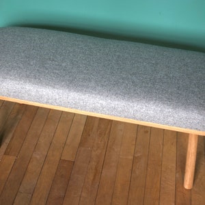 Könnte beinhalten: Eine graue gepolsterte Bank mit einer abgerundeten ovalen Oberseite und Holzbeinen. Die Bank steht auf einem Holzboden.