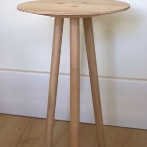 May include: A small, light-colored wooden side table with a round top and three cylindrical legs. The table is made of natural wood and stands against a white wall. The table's top has a diameter of approximately 18 inches.