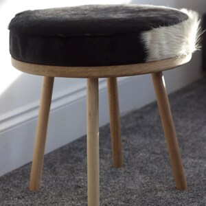 May include: A round stool with a black and white cowhide seat. The seat is supported by a light-colored wooden frame and three angled legs. The stool is a decorative piece of furniture.