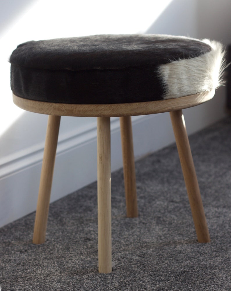May include: A round stool with a black and white cowhide seat. The seat is supported by a light-colored wooden frame and three angled legs. The stool is a decorative piece of furniture.