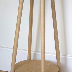 May include: A wooden plant stand with a round top and three legs. The stand is made of light-colored wood and has a natural finish.