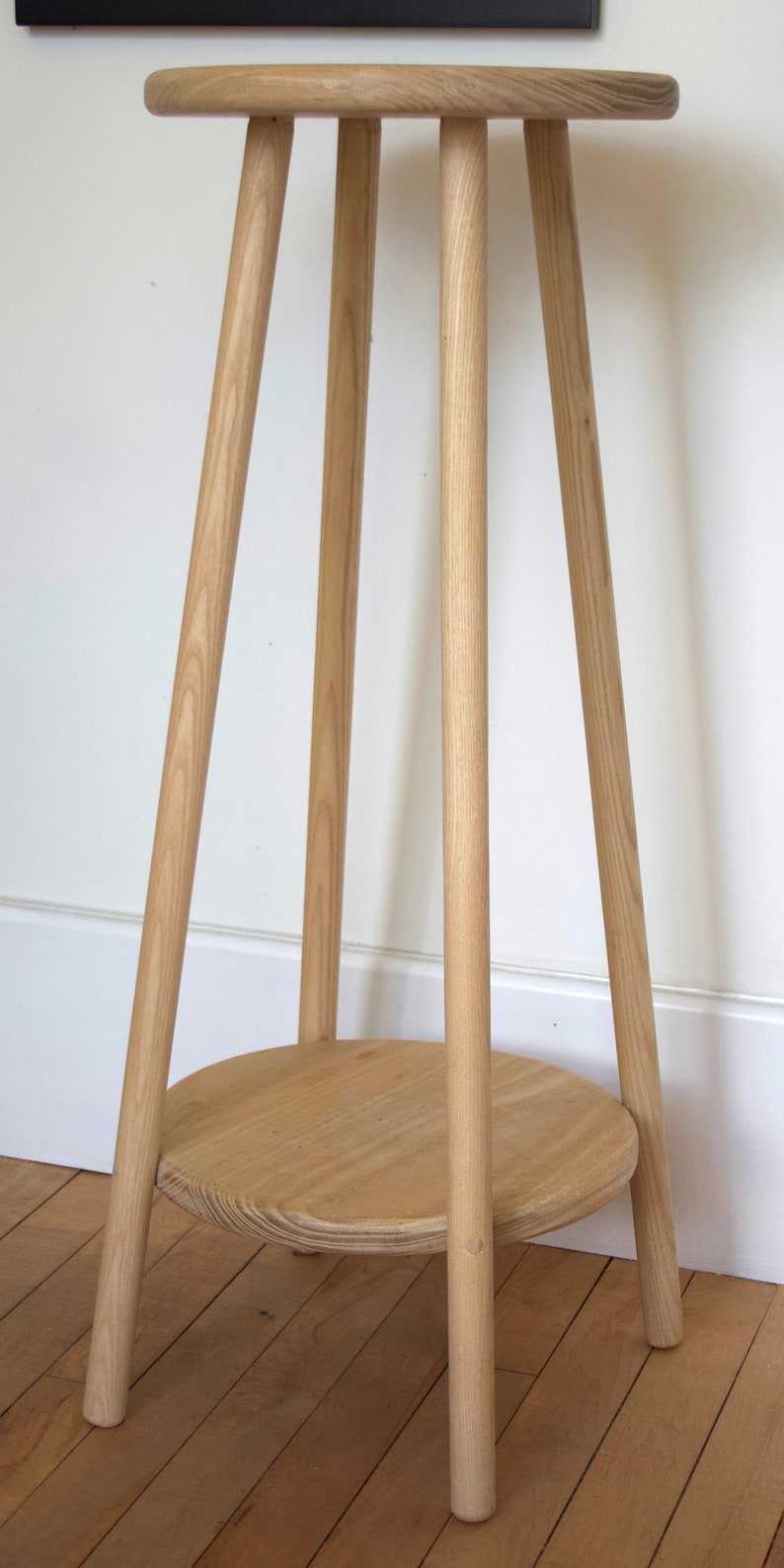 May include: A wooden plant stand with a round top and three legs. The stand is made of light-colored wood and has a natural finish.