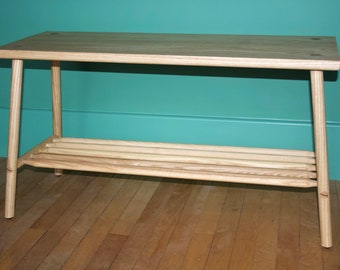 May include: A light-colored wooden bench featuring a rectangular top surface and four tapered legs. The bench includes a lower storage shelf constructed from several parallel wooden rods, providing an open and airy design structure.