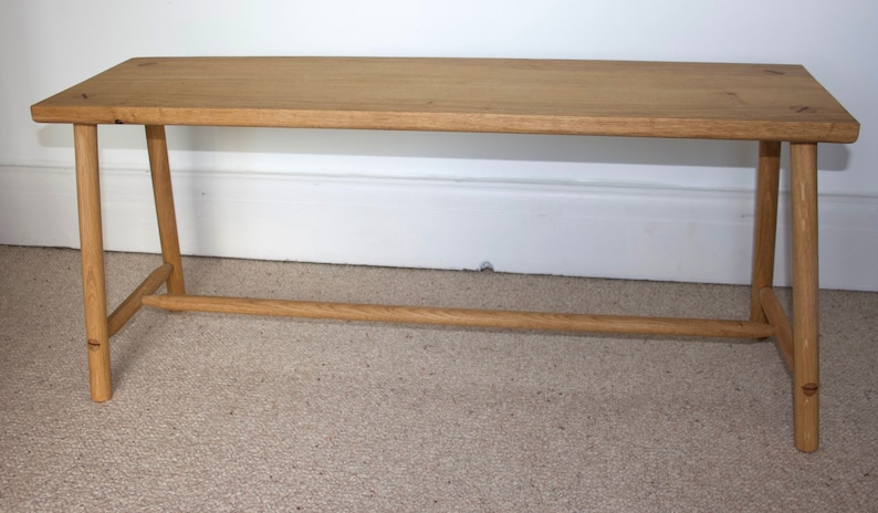 May include: A rectangular wooden bench featuring a light brown natural wood grain finish. The bench has four straight, cylindrical legs connected by a horizontal support stretcher bar running between the legs on each side for stability.