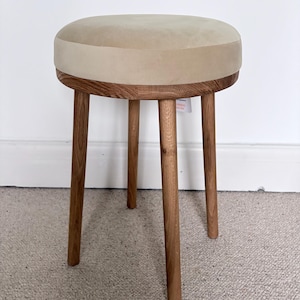 May include: A round stool featuring a beige upholstered seat cushion and a wooden frame with three cylindrical legs. The wood grain is visible on the legs and the circular base supporting the seat, which has a smooth, light-colored fabric top.