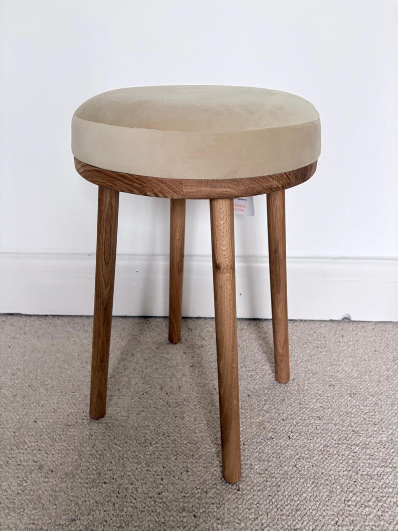 May include: A round stool featuring a beige upholstered seat cushion and a wooden frame with three cylindrical legs. The wood grain is visible on the legs and the circular base supporting the seat, which has a smooth, light-colored fabric top.