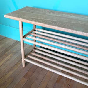 May include: A light-colored wooden bench with a slatted lower shelf. The bench has a rectangular top and four rounded legs. The shelf is made of multiple horizontal wooden slats. The bench is made of light-colored wood.