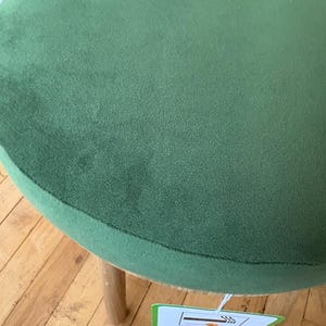 May include: A round stool featuring a soft, olive green velvet seat cushion and light-colored wooden legs. A white rectangular tag is attached to the underside of the seat, displaying a graphic icon of a cigarette and the word RESISTANT.