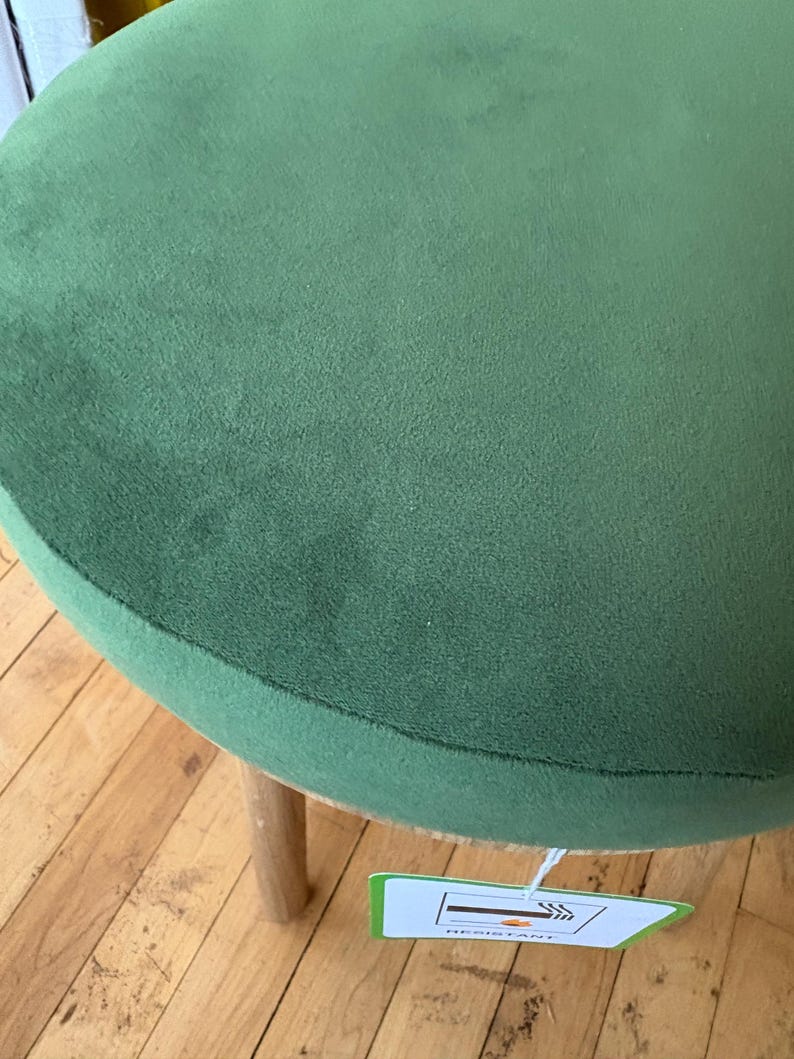 May include: A round stool featuring a soft, olive green velvet seat cushion and light-colored wooden legs. A white rectangular tag is attached to the underside of the seat, displaying a graphic icon of a cigarette and the word RESISTANT.