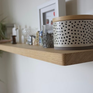 May include: A light-coloured wooden floating shelf mounted on a white wall. The shelf holds a cylindrical container with a cream-coloured base and black polka dots, topped with a wooden lid, alongside various small decorative items and a green plant.
