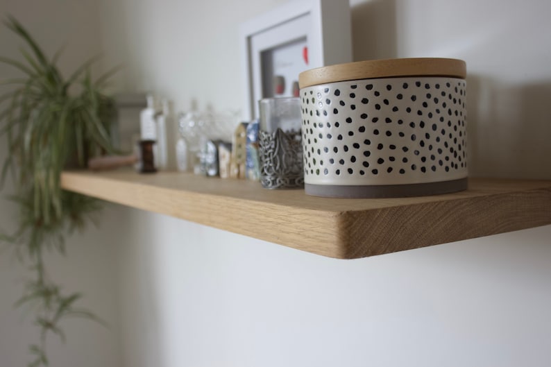 May include: A light-coloured wooden floating shelf mounted on a white wall. The shelf holds a cylindrical container with a cream-coloured base and black polka dots, topped with a wooden lid, alongside various small decorative items and a green plant.