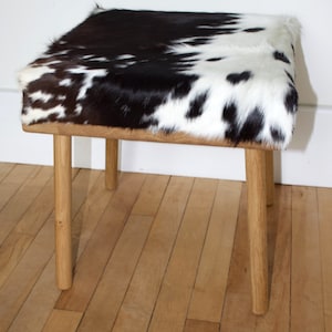 Könnte beinhalten: Ein quadratischer Hocker mit einer schwarzen und weißen Kuhfell-Oberfläche und vier Holzbeinen. Der Hocker steht auf einem Holzboden.