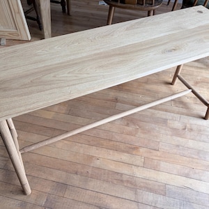 May include: A rectangular light-colored wooden bench featuring a smooth grain surface, four slender cylindrical legs, and a horizontal support stretcher bar. The tabletop is secured with visible circular wooden dowel joinery at each corner.