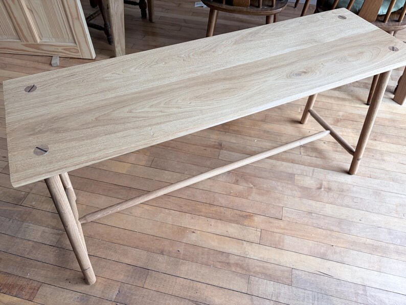 May include: A rectangular light-colored wooden bench featuring a smooth grain surface, four slender cylindrical legs, and a horizontal support stretcher bar. The tabletop is secured with visible circular wooden dowel joinery at each corner.