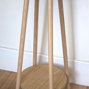 May include: A light wood plant stand with a round top and a lower shelf. The stand has three legs and is made of natural wood.