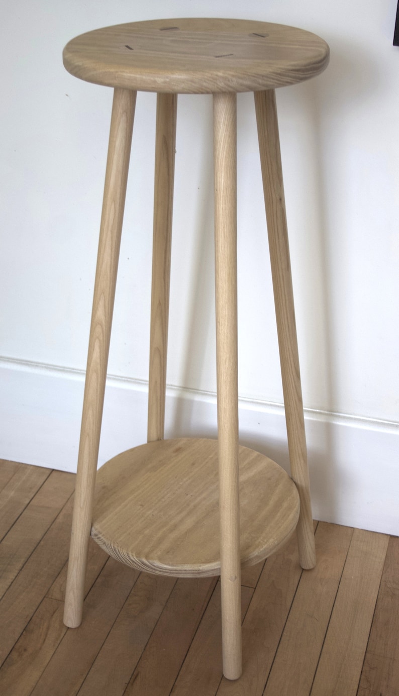 May include: A light wood plant stand with a round top and a lower shelf. The stand has three legs and is made of natural wood.