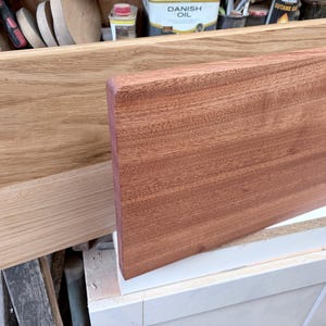 May include: Two rectangular wooden boards are displayed. The board in the back is a light tan oak with a visible grain pattern. The board in the front is a reddish-brown mahogany with a smooth, uniform surface and rounded edges.