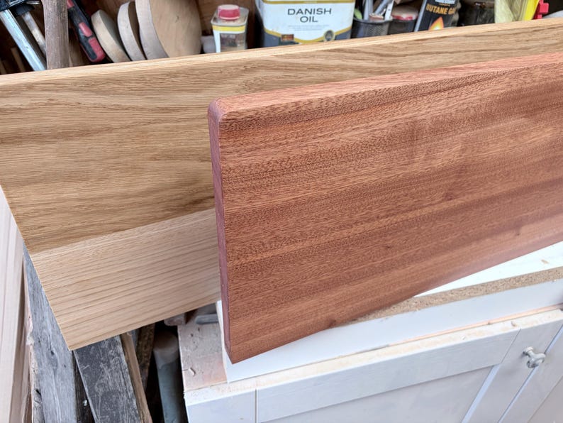 May include: Two rectangular wooden boards are displayed. The board in the back is a light tan oak with a visible grain pattern. The board in the front is a reddish-brown mahogany with a smooth, uniform surface and rounded edges.
