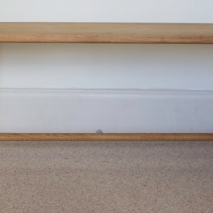 May include: A light brown wooden bench featuring a rectangular seat with rounded corners and four splayed legs. The legs are connected by a horizontal support bar on each side, showcasing a natural wood grain finish and visible joinery details.