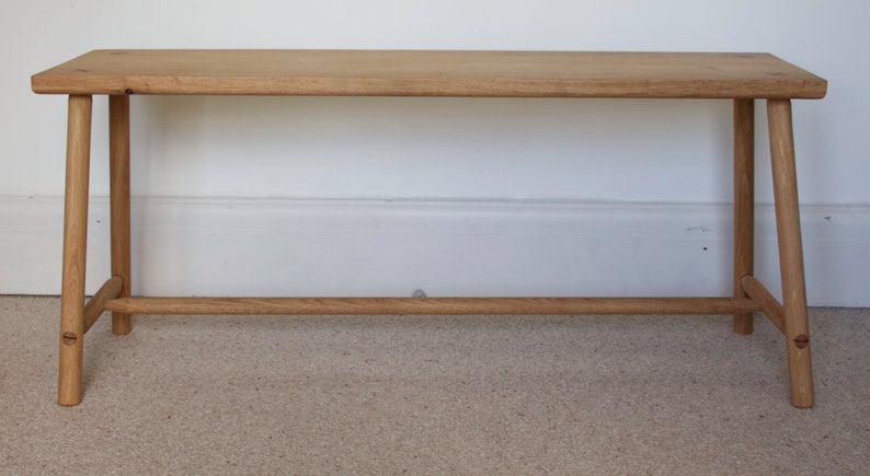 May include: A light brown wooden bench featuring a rectangular seat with rounded corners and four splayed legs. The legs are connected by a horizontal support bar on each side, showcasing a natural wood grain finish and visible joinery details.