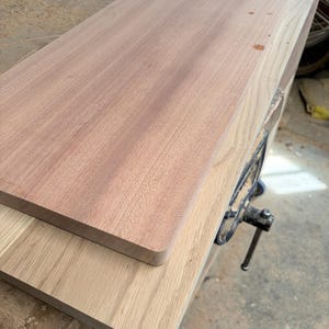 May include: Two rectangular wooden boards stacked on top of each other. The top board is a reddish-brown wood with a smooth, straight grain. The bottom board is a lighter, pale tan wood with a visible, linear grain pattern.