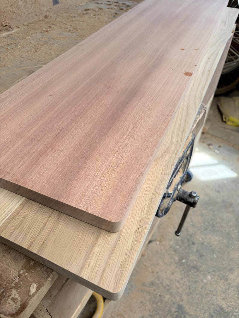 May include: Two rectangular wooden boards stacked on top of each other. The top board is a reddish-brown wood with a smooth, straight grain. The bottom board is a lighter, pale tan wood with a visible, linear grain pattern.
