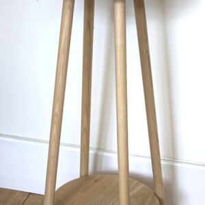 May include: A light wood plant stand with a round top and a lower shelf. The stand has four legs and is made of natural wood. A gray planter with a succulent plant is sitting on the top shelf.